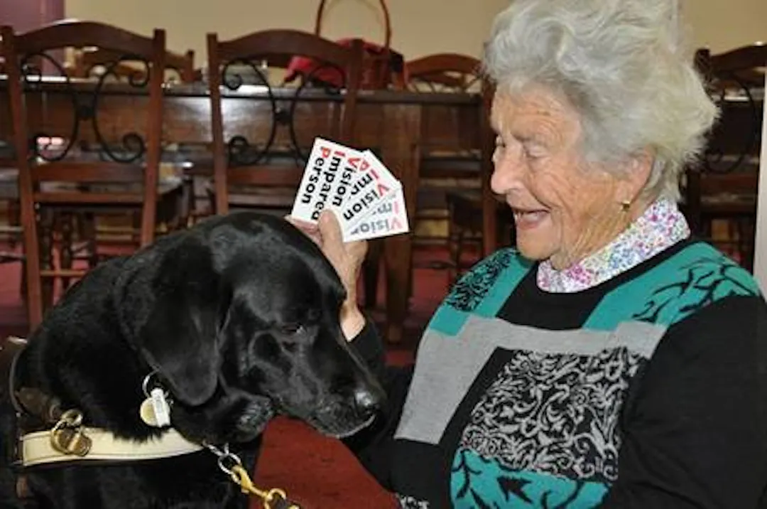 Black-and-white photograph of a person seated on a chair indoors, holding the handle of a guide dog lying in the foreground